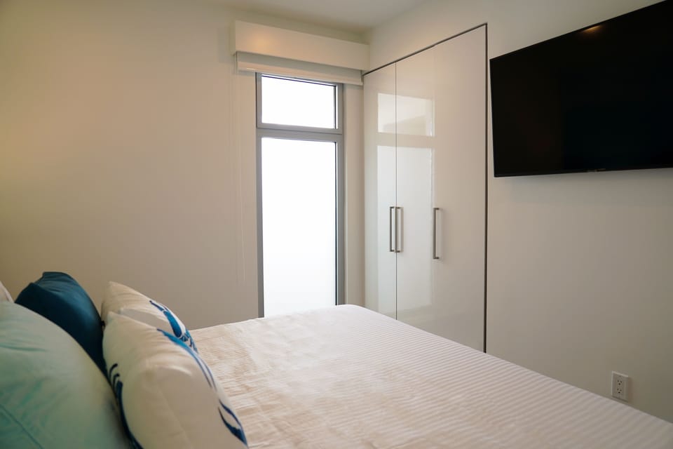 Crisp white linens and plush pillows invite you to sink in and unwind. Bright windows bring in natural light, while the sleek wardrobe and wall-mounted TV add modern comfort to your restful retreat.