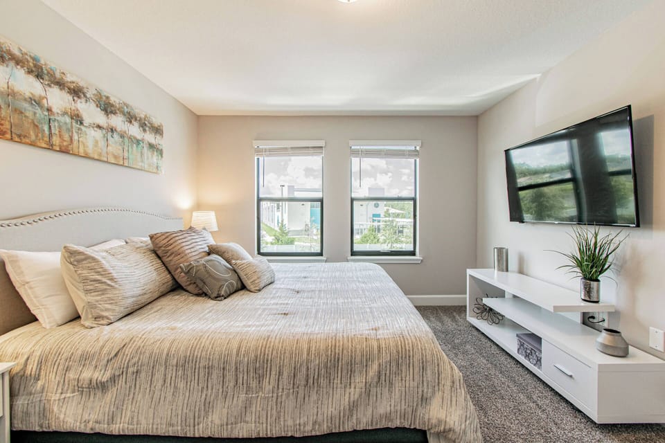 Awake to natural light in this cozy bedroom featuring soft gray tones and plush bedding, inviting relaxation. Contemporary white furniture and tasteful artwork enhance the calm ambiance, perfect for unwinding after a long day.