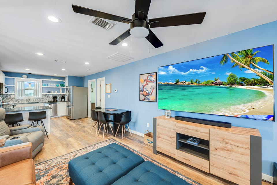 Unwind in your open-concept living space featuring a large TV, modern kitchen, and tropical blue tones that create a breezy coastal atmosphere.