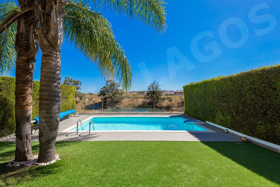 Beautifully maintained private garden at Casa Vista Lago, Lagos, with a manicured green lawn, mature palm tree providing natural shade, a sparkling swimming pool with stone surround, and tall hedging ensuring complete privacy for guests