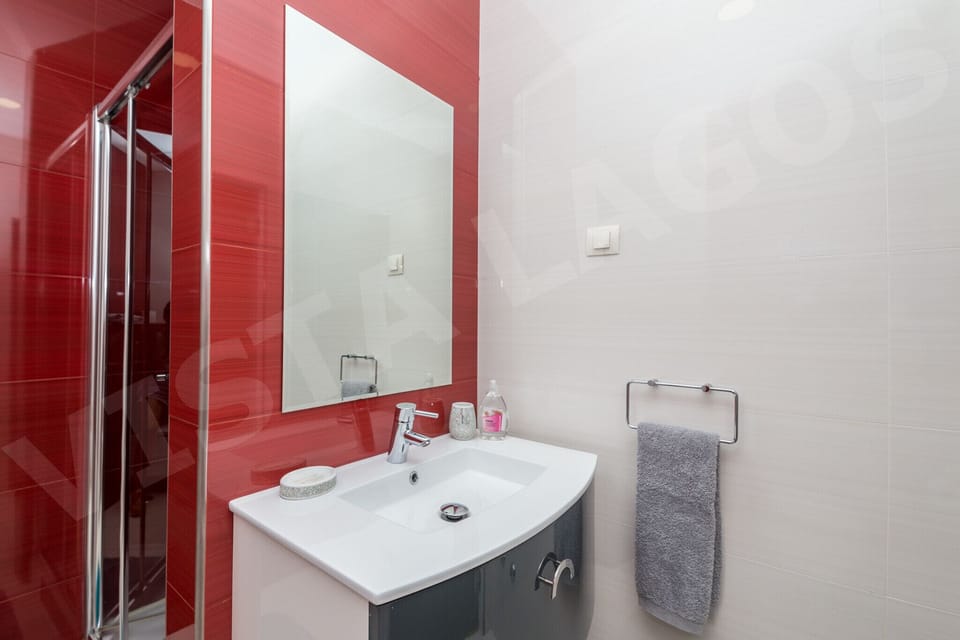 Modern en-suite bathroom in Casa Vista Lago, Lagos, featuring bold red tiled walls, a spacious glass-enclosed walk-in shower, a white vanity unit with basin and large wall mirror, chrome fittings, and fresh towels provided for guests
