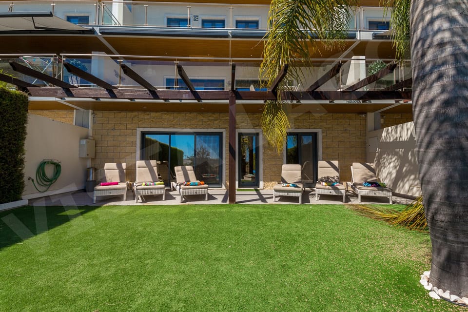 Rear exterior view of Casa Vista Lago showing the covered pergola terrace with sun loungers on a manicured green lawn, a 4-bedroom holiday townhouse in Lagos, Algarve, with large glass doors leading to the garden