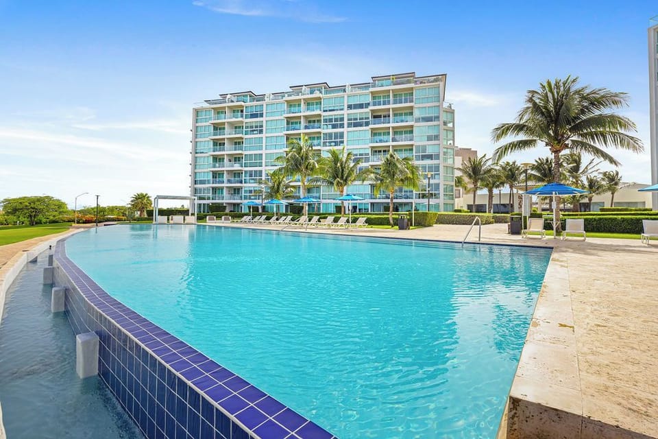 Modern resort building with expansive swimming pool surrounded by tropical palm trees under clear blue skies.