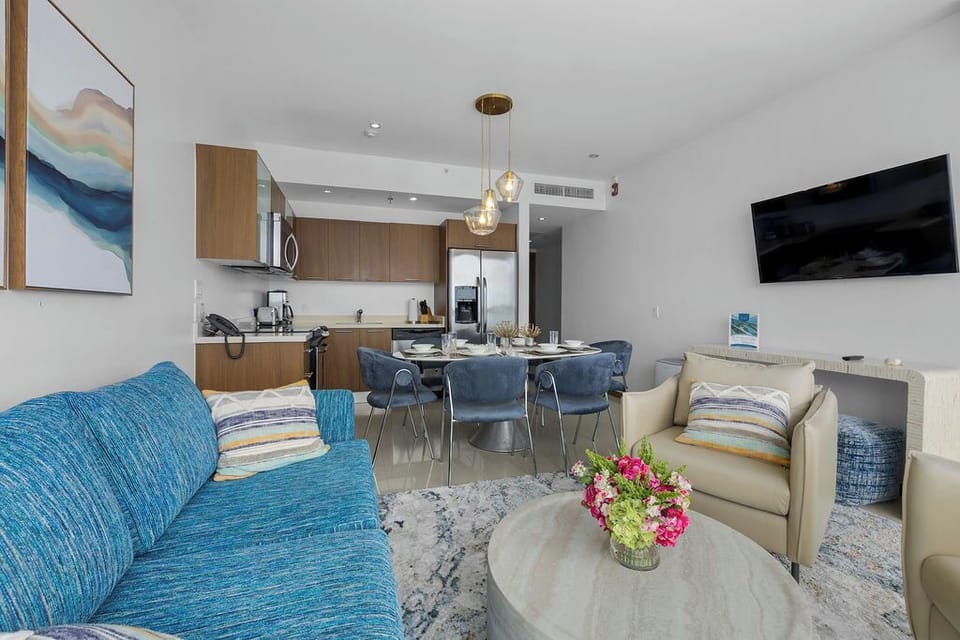 Your bright open space flows seamlessly from cozy sofa to dining table to modern kitchen, perfect for gathering with loved ones while coastal artwork adds pops of calming color.