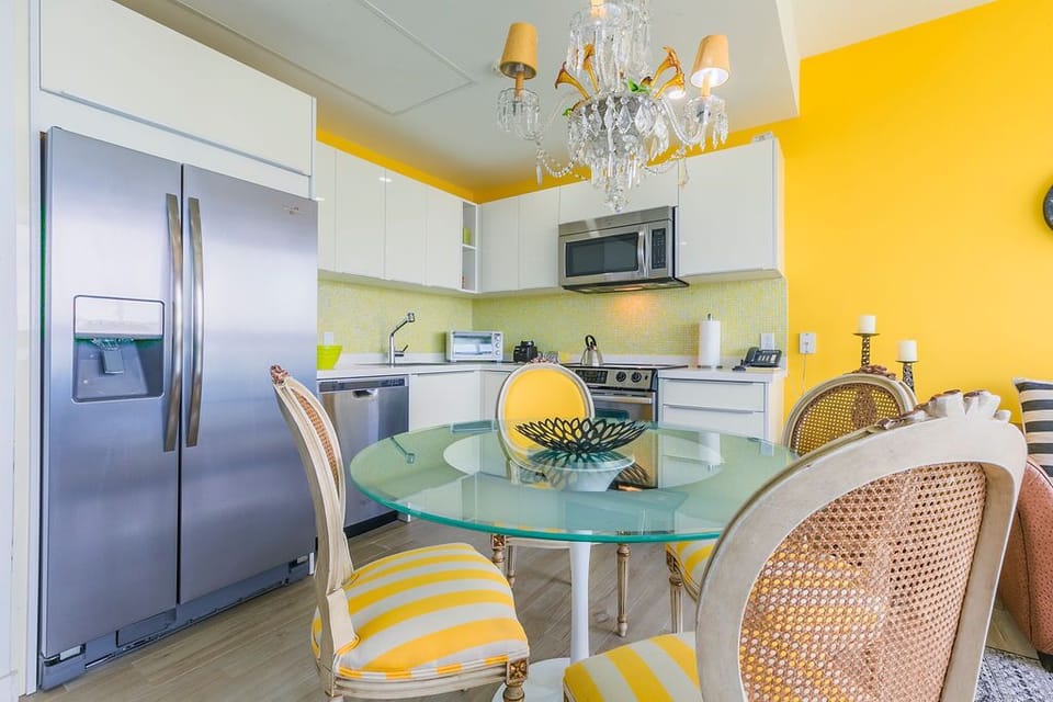Your bright modern kitchen welcomes you with cheerful yellow walls, gleaming stainless appliances, and a chic glass dining table beneath an elegant chandelier.