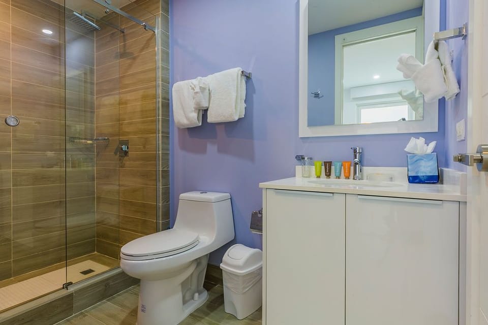 Your modern bathroom features a sleek glass-enclosed shower with warm wood-look tile, crisp white vanity, and soothing periwinkle walls—fresh towels ready for your arrival.
