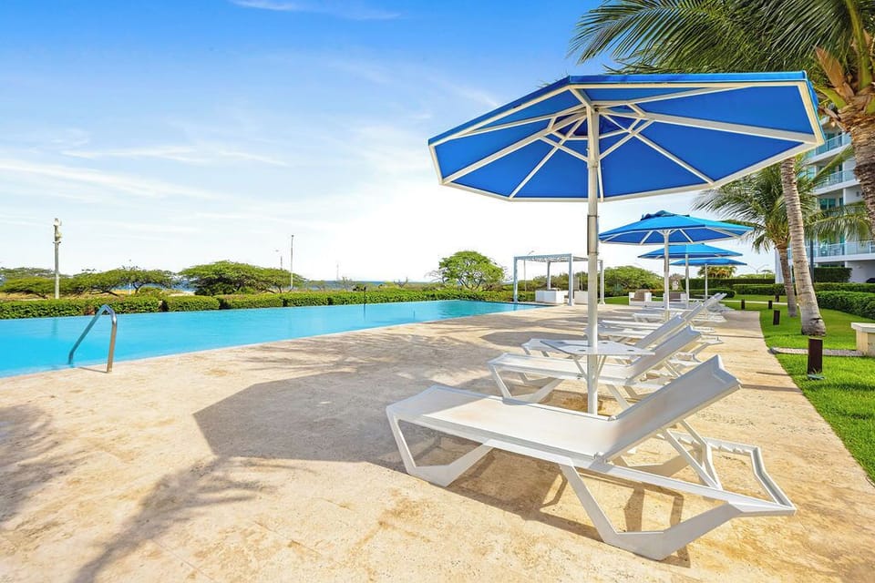 Pristine pool deck with azure water and loungers under blue umbrellas, surrounded by swaying palms and tropical gardens.