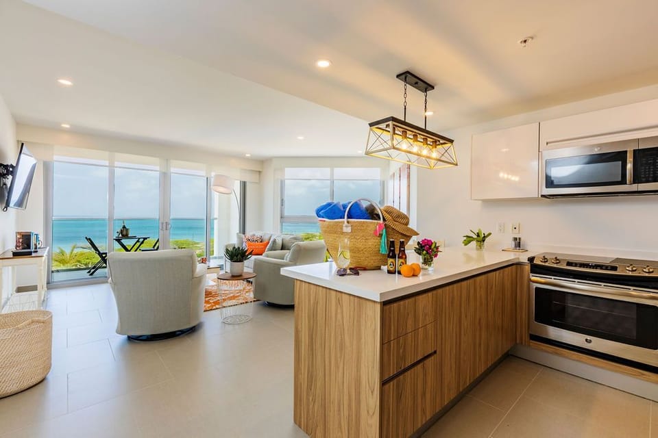 Step into your bright, modern haven where the kitchen flows seamlessly into the living space. Through floor-to-ceiling windows, turquoise waters and coastal greenery frame your days, creating a serene backdrop for cooking, relaxing, and savoring every mome