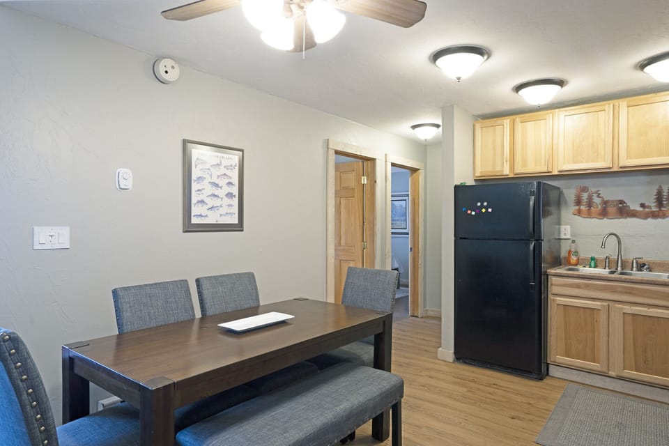 A small dining area with a dark wood table and four upholstered chairs. Behind, there is a kitchen with wooden cabinets, a black refrigerator, and a countertop with a sink. Ceiling lights are on.