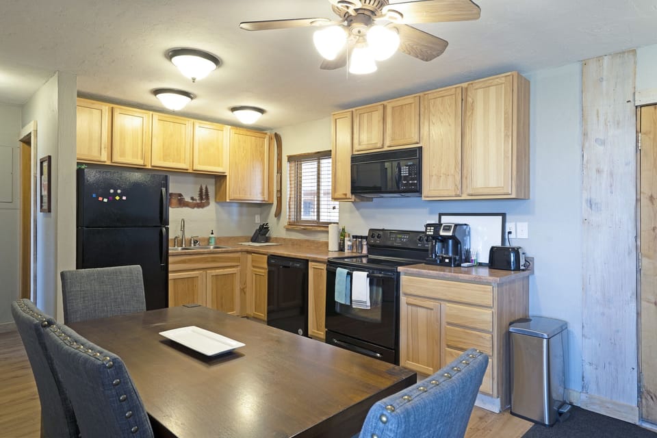 A kitchen with wooden cabinets, a black refrigerator, black stove and microwave, a sink, and various kitchen appliances on the countertop. A dining table with chairs is in the foreground.