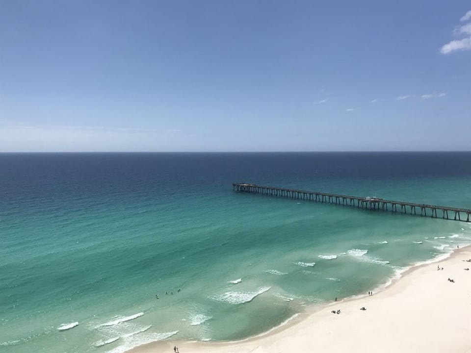 Stunning emerald waters and pristine white sand stretch along the coastline, featuring a wooden pier extending into crystal-clear Gulf waters.
