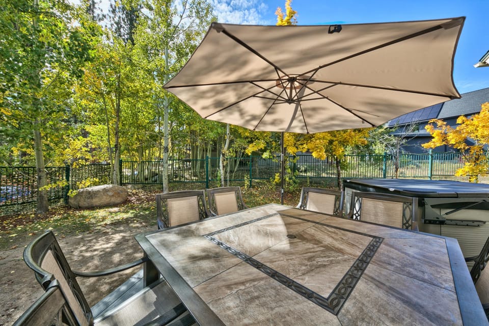 Outdoor dining area with stone tile table, metal-framed chairs, large beige umbrella, and fenced yard with trees.