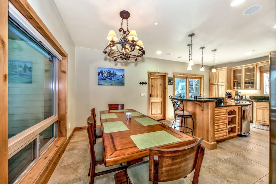 Dining area with a wooden table set for six beneath a metal chandelier, adjacent to a kitchen island with granite counters.