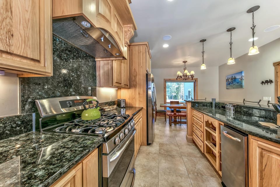 Galley kitchen with polished green granite counters, natural wood cabinetry, stainless steel appliances, and tile flooring.