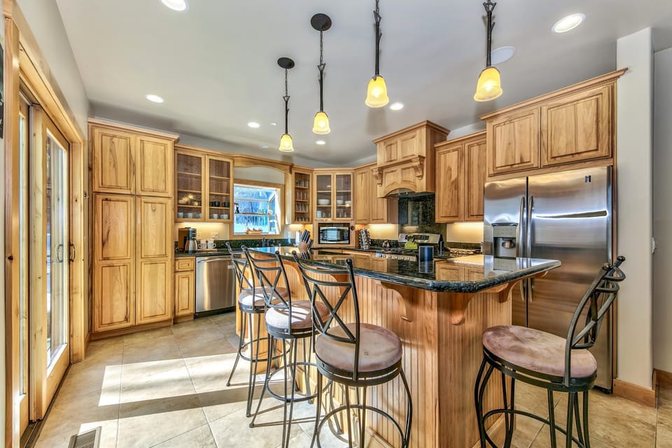 Sunlight strikes tile floors beneath a wood-paneled island with black granite counters and metal barstools.