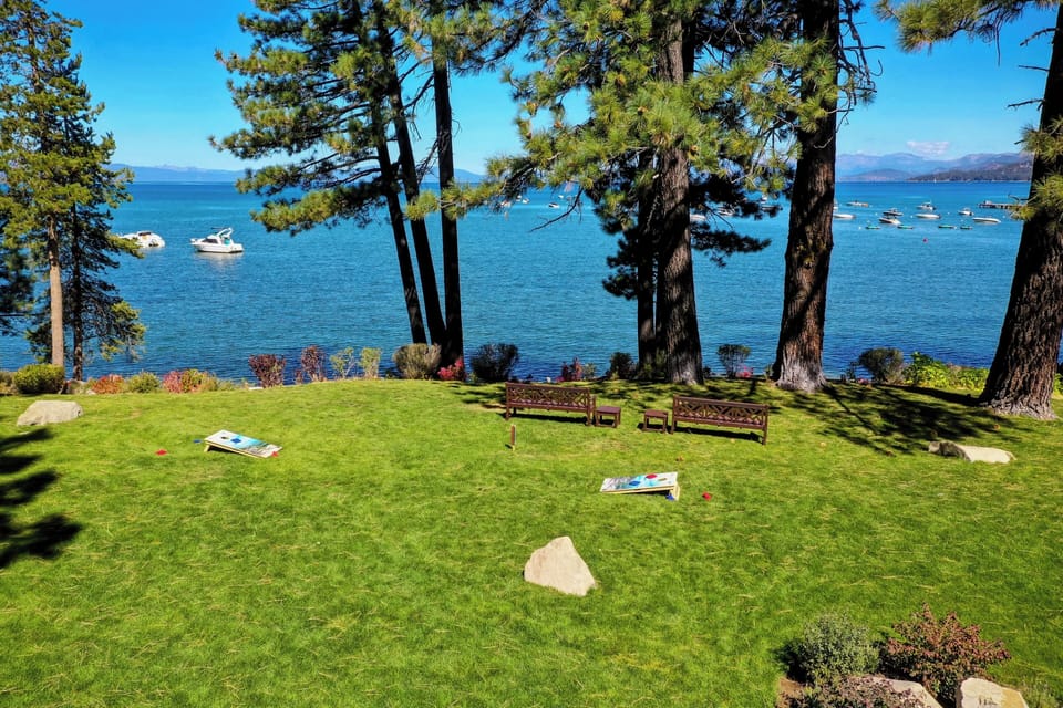 Sierra Shores community lakeside lawn framed by towering pines offers stunning views of blue waters, distant mountains, and anchored boats, with cornhole boards ready for sunny afternoon games.