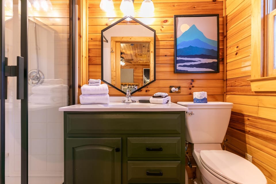 Cedar-wrapped and cozy, this bathroom at Riverbend Retreat pairs a forest-green vanity with mountain artwork and a roomy glass shower.