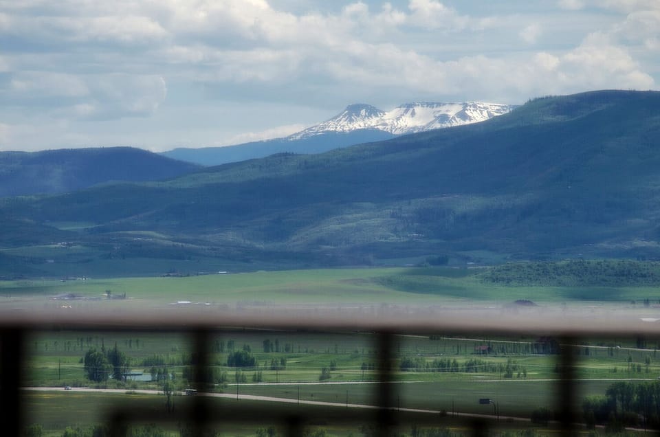 Mountain landscape with snow-capped peaks rising above rolling green valleys and farmland under a cloudy sky.