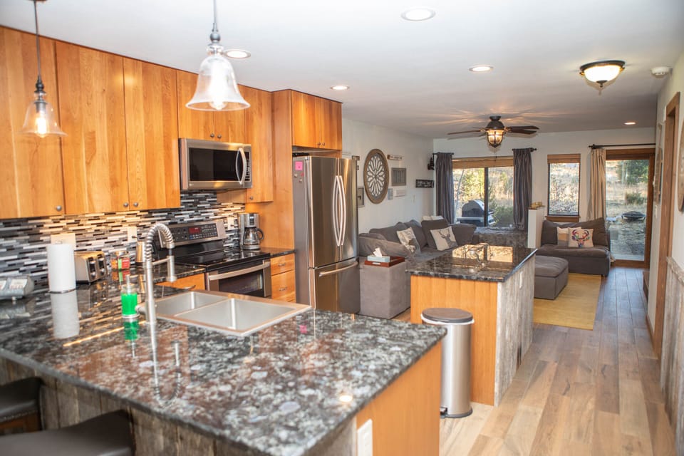 Modern kitchen with wooden cabinets, stainless steel appliances, and granite countertops, extending into an open living area with gray sofas, wooden flooring, and large windows.