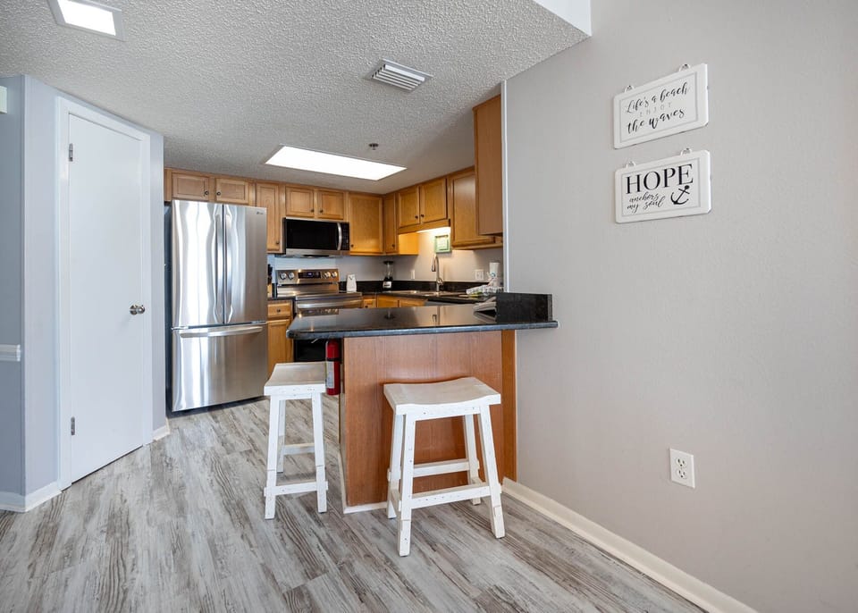 The kitchen features warm wood cabinetry, a sleek black countertop, and a breakfast bar with white stools