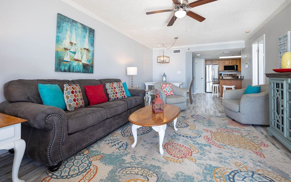 The living room features a plush gray sofa adorned with colorful pillows, a wooden coffee table, and a vibrant area rug