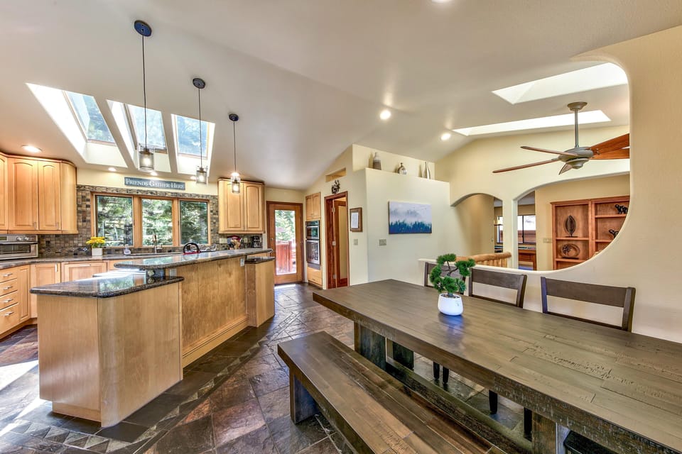 Dining area with rustic wood table and bench beneath skylights, adjacent to kitchen featuring granite countertops and light wood cabinetry.