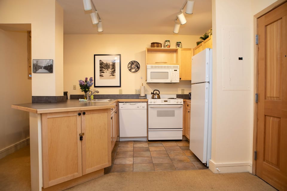 A small kitchen with light wood cabinets, white appliances, and track lighting. The kitchen features a stove, microwave, refrigerator, dishwasher, and a small countertop with a sink and flowers.