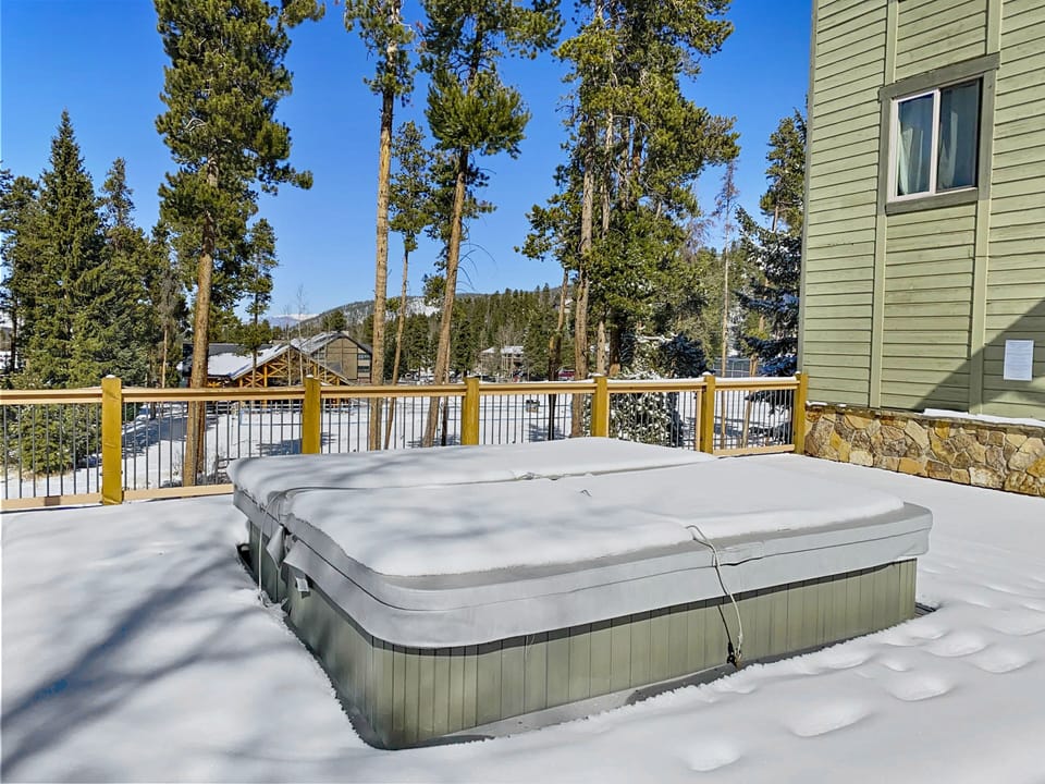 A closed hot tub on a snowy deck next to a green building, surrounded by tall trees and a snowy landscape.