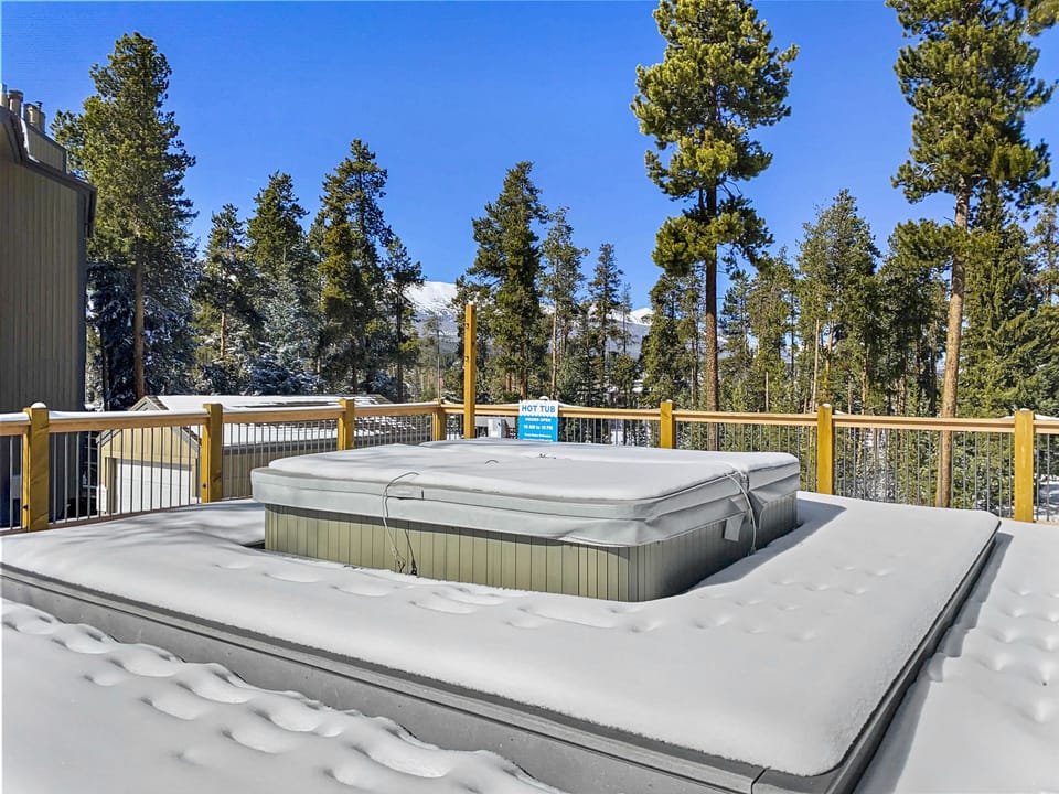 Snow-covered hot tub on a rooftop deck with a wooden railing, surrounded by tall trees and a clear blue sky in the background.