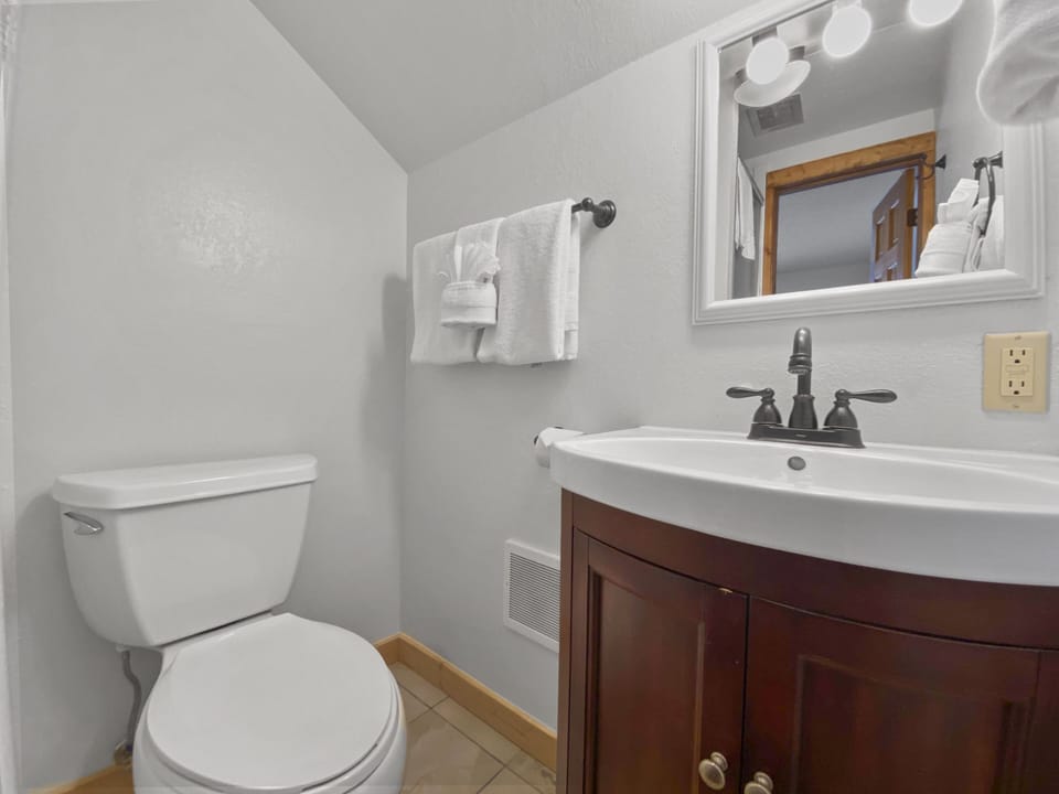 Small bathroom with a white toilet, dark wood vanity with white sink, wall-mounted mirror, towel rack with folded towels, and light fixture above the mirror. Walls are painted white.
