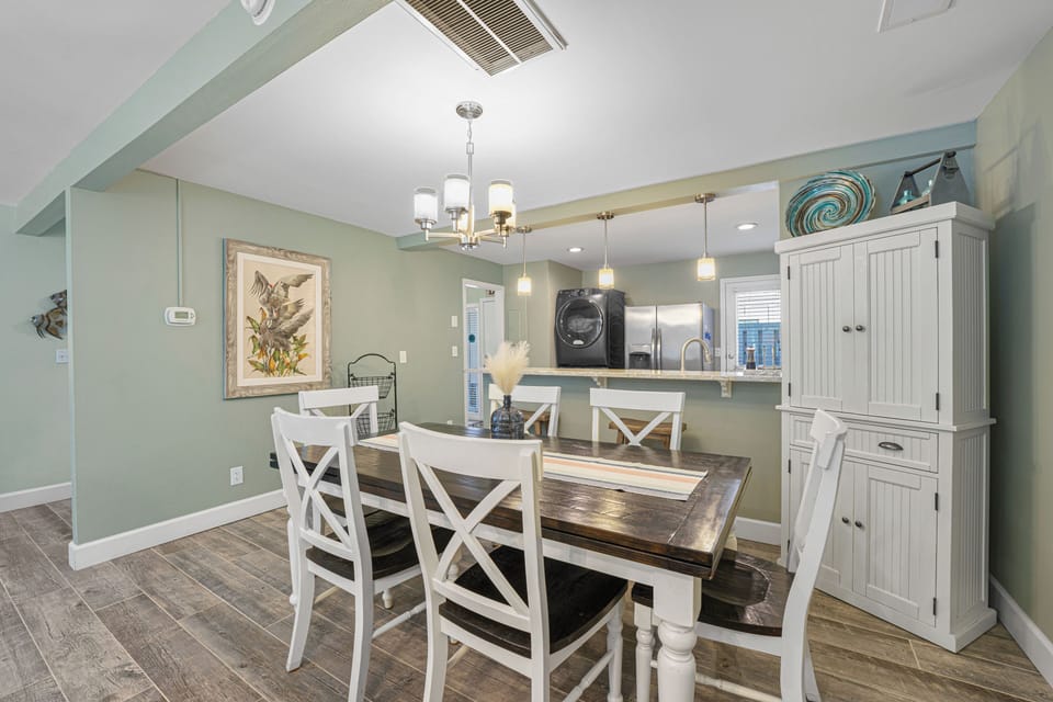 A bright and coastal dining area featuring a farmhouse-style table, soft sea-glass walls, and open flow to the kitchen