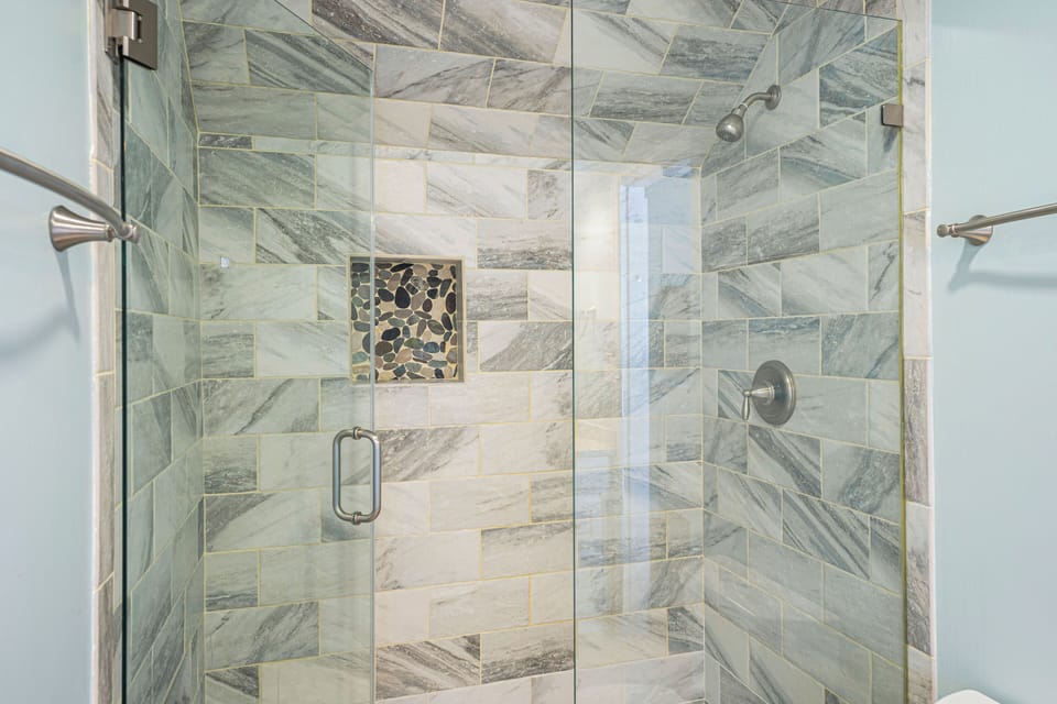 Elegant walk-in shower featuring a glass enclosure, marble-style tiles, and a decorative stone accent niche for a modern coastal touch.