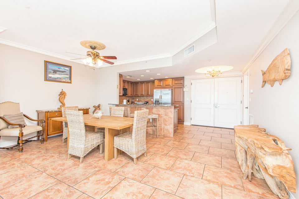 Step into a bright, coastal-inspired living space where warm terracotta tiles, wicker seating, and beach-themed décor create a relaxing retreat. The open layout flows seamlessly into your fully equipped kitchen, perfect for meals together.