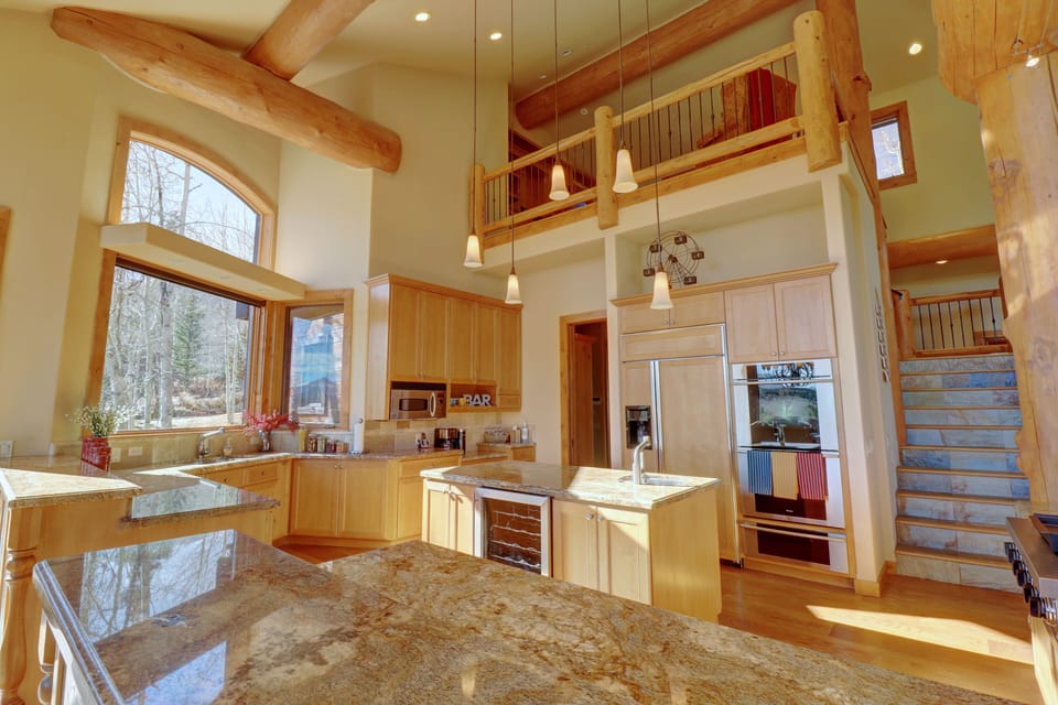 A spacious kitchen with wooden cabinets, granite countertops, stainless steel appliances, and a high ceiling with exposed beams. Stairs with a wooden rail and a balcony are visible in the background.