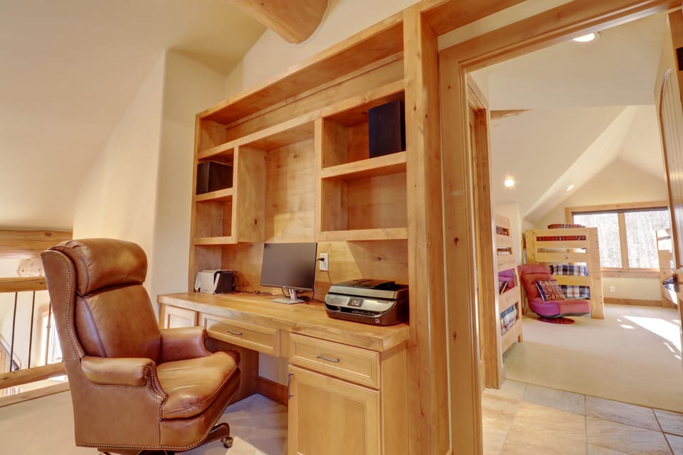 A home office with a wooden desk, leather chair, computer, and printer. In the background, a hallway leads to a room with bunk beds and a window with natural light.
