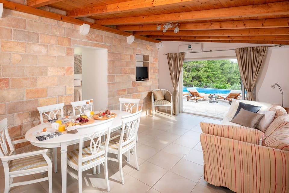 The interior of the living room of the villa Zlatni Rat in Bol with white living room furniture, a living room with a sofa, an armchair and a TV, and a view of the courtyard with a swimming pool.