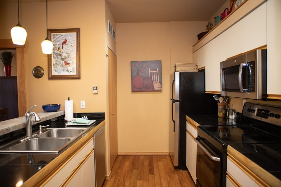 A modern kitchen with black appliances, light wood cabinetry, a double sink, and a black countertop. Artwork hangs on beige walls, and pendant lights illuminate the space.