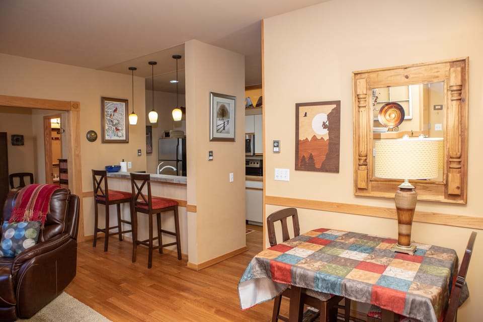 A cozy dining area with a colorful tablecloth, a lamp on a side table, and a pass-through to a kitchen with bar stools and pendant lights. There's a patterned chair and framed artwork on the walls.