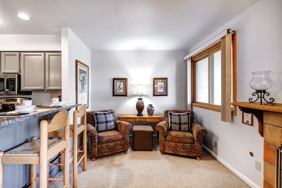 The living room with 2 upholstered chairs, throw pillows, table with lamp and vase, ottoman and 2 pictures on the wall.