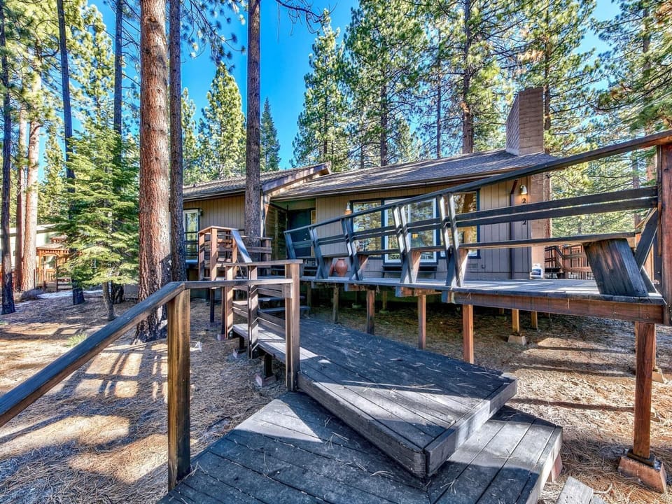 Forest cabin with elevated deck and stairs, surrounded by towering pine trees and mountain wilderness.
