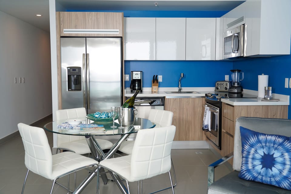 You'll love preparing meals in this modern kitchen with sleek appliances, vibrant blue accents, and a stylish glass dining table—perfect for enjoying morning coffee or evening dinners together.