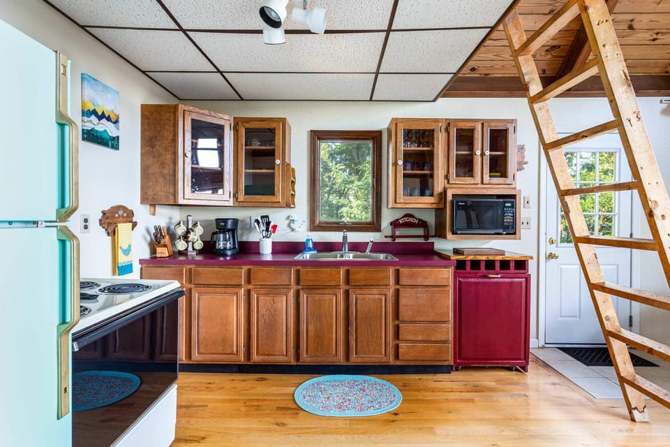 Cozy kitchen with wooden cabinets, a window view, and a rustic ladder leading to a loft.