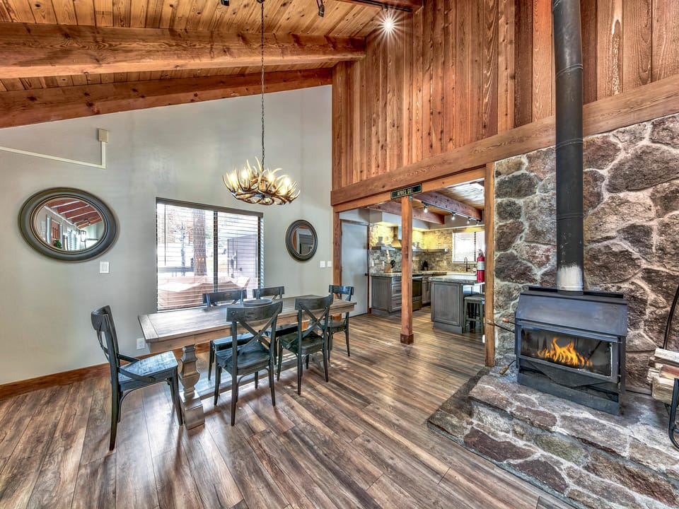 Dining area with antler chandelier, rustic wood table, black chairs, and stone fireplace with wood stove insert.