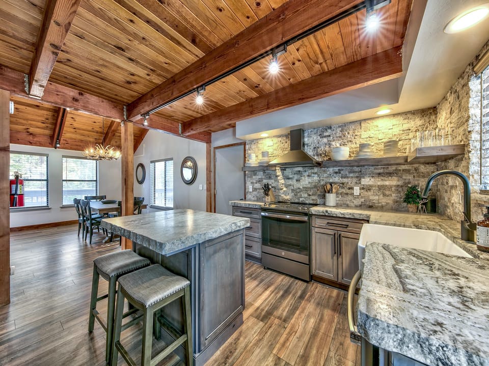 Granite island countertop with two cushioned stools, wood-plank floors, and exposed wood ceiling beams in kitchen.