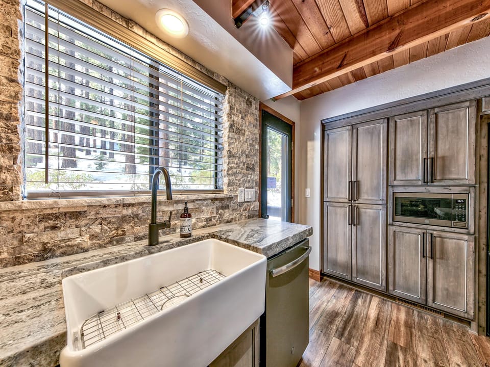Farmhouse sink with a brushed black faucet sits beneath a large window with stone tile backsplash and wood-plank ceiling.
