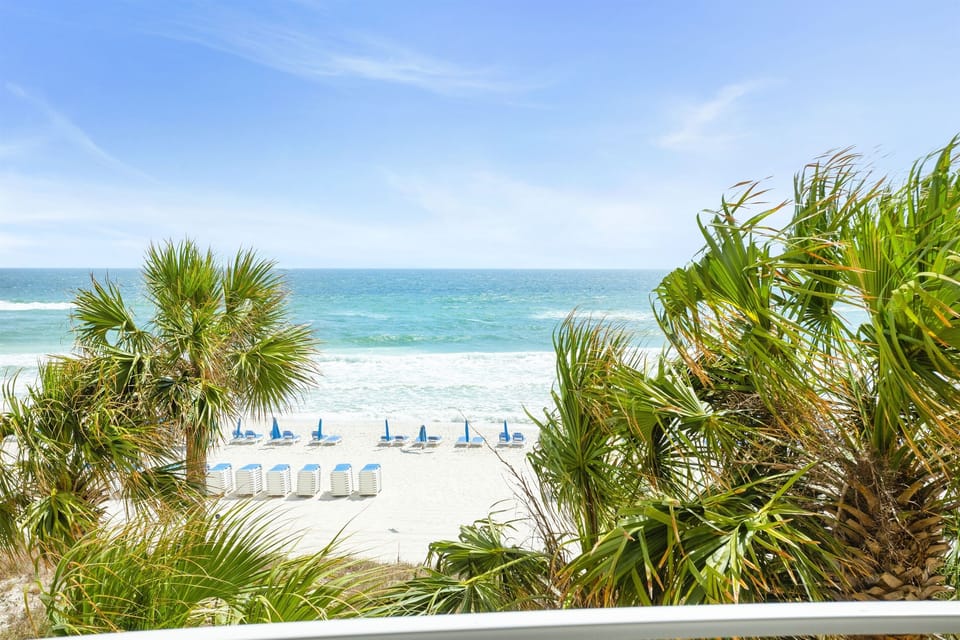 Pristine white sand beach framed by tropical palms with turquoise waters and beach umbrellas dotting the shoreline.