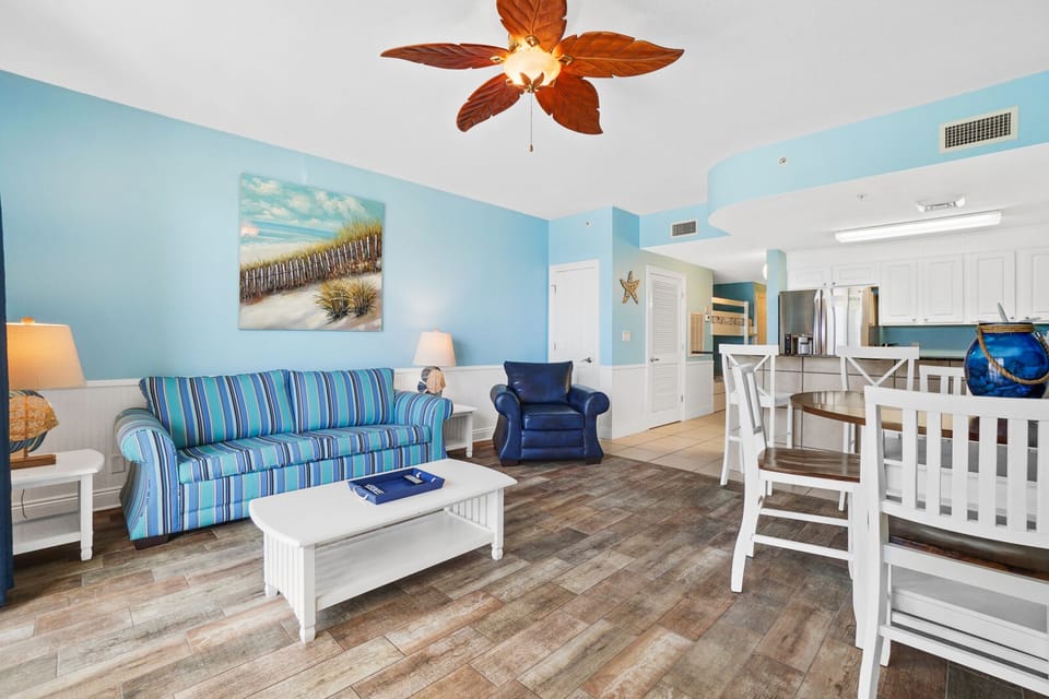 Relax in your bright, coastal-themed living space featuring comfy striped seating, tropical ceiling fan, and open layout connecting to your fully equipped kitchen.