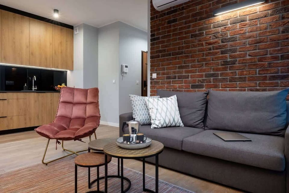 Cohesive combination of living room and kitchenette. A brick wall is visible behind the grey sofa.