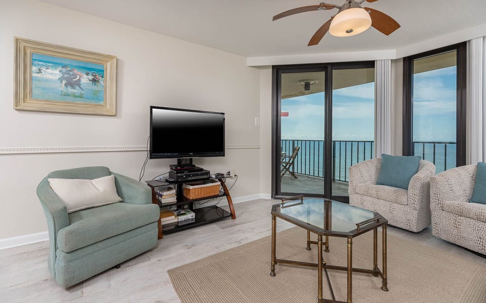 Living area features a light blue armchair, two white sofas, a glass coffee table, and the balcony with a beach front view
