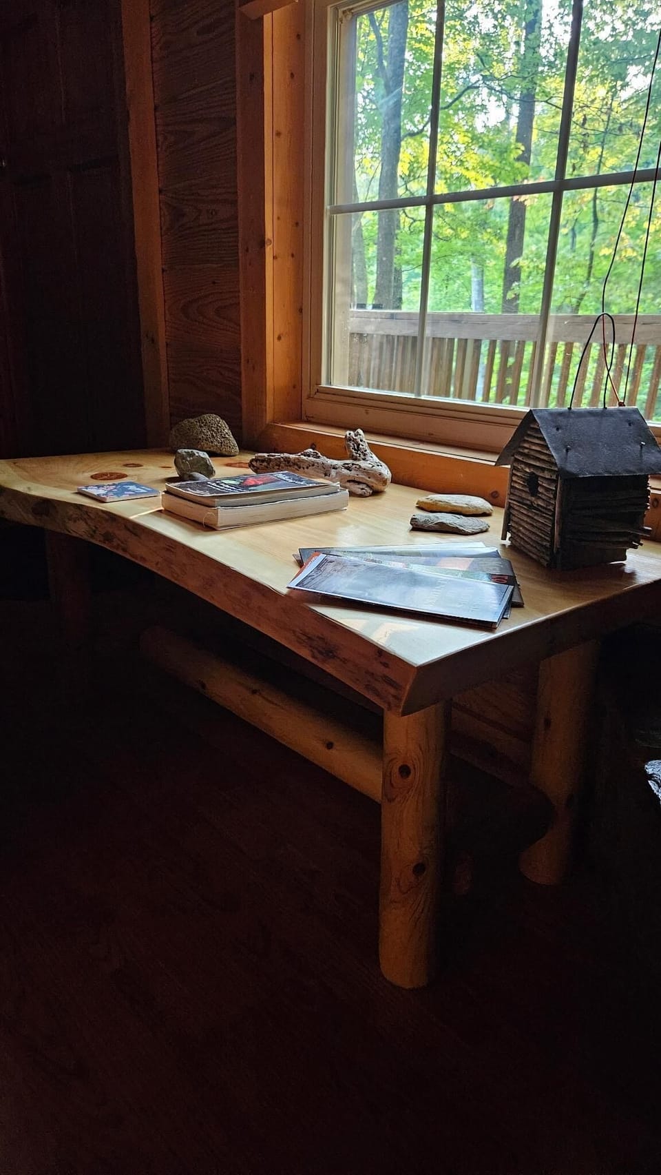 Live-edge wooden desk beneath a window, displaying books, driftwood, stones, and a rustic birdhouse.
